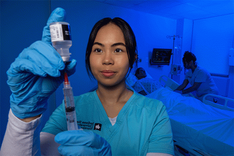 Tan-skinned female student with dark black hair and wearing blue scrubs looks closely at a hypodermic needle while drawing an injection, with another nurse working on a patient simulator in a hospital bed in the background. The room has a blue tint.   